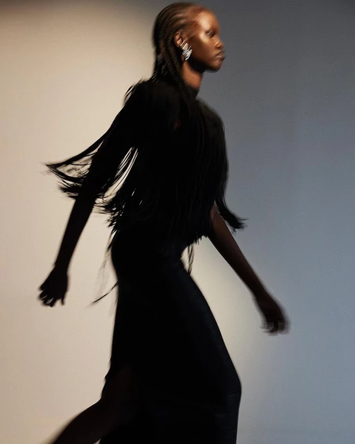 A slender woman in motion wearing a fringe-detailed black dress and large earrings against a soft, gradated background, fashion photography.