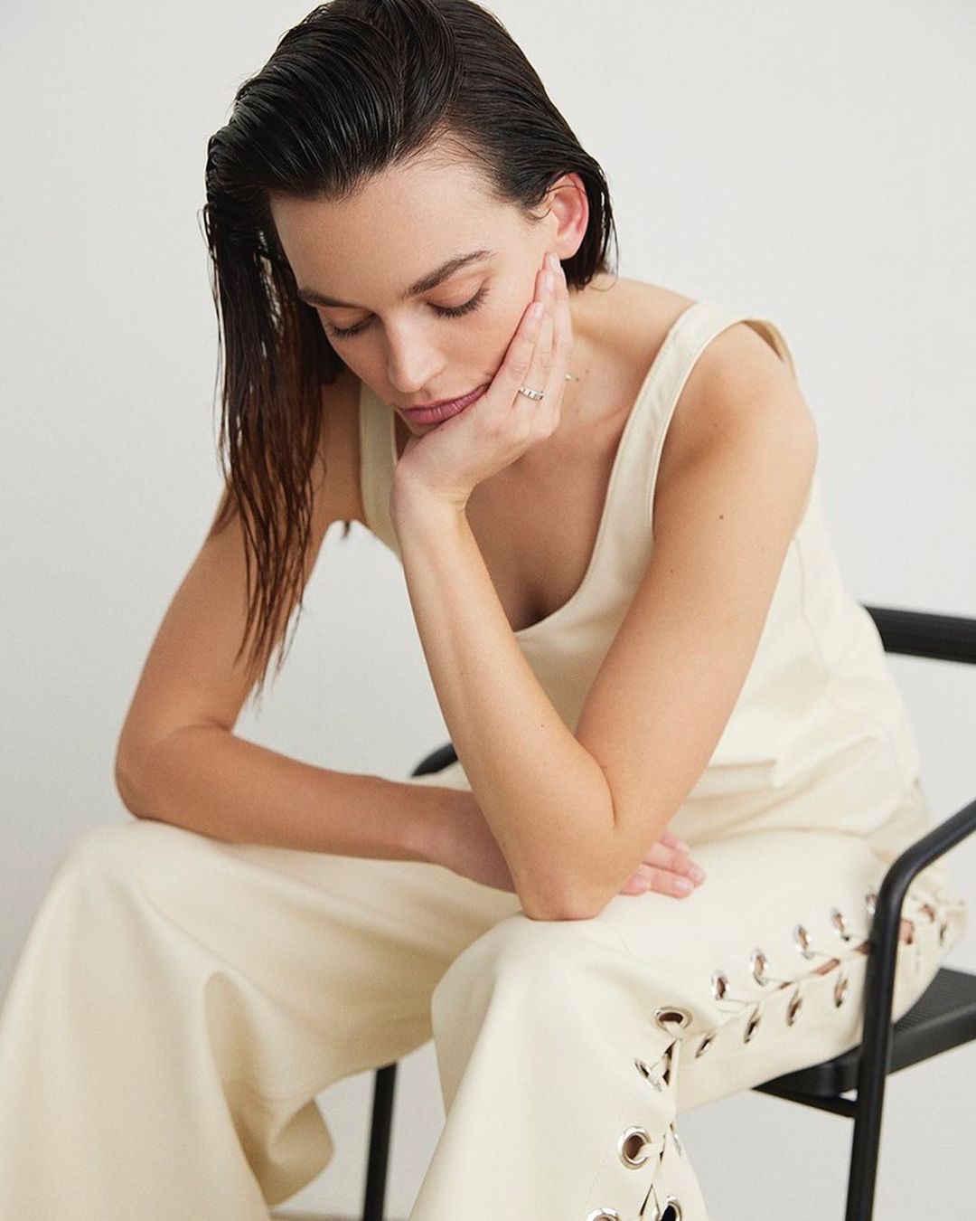 Woman in cream top and pants seated on chair with head resting on hand looking down in a minimalist indoor studio setting.