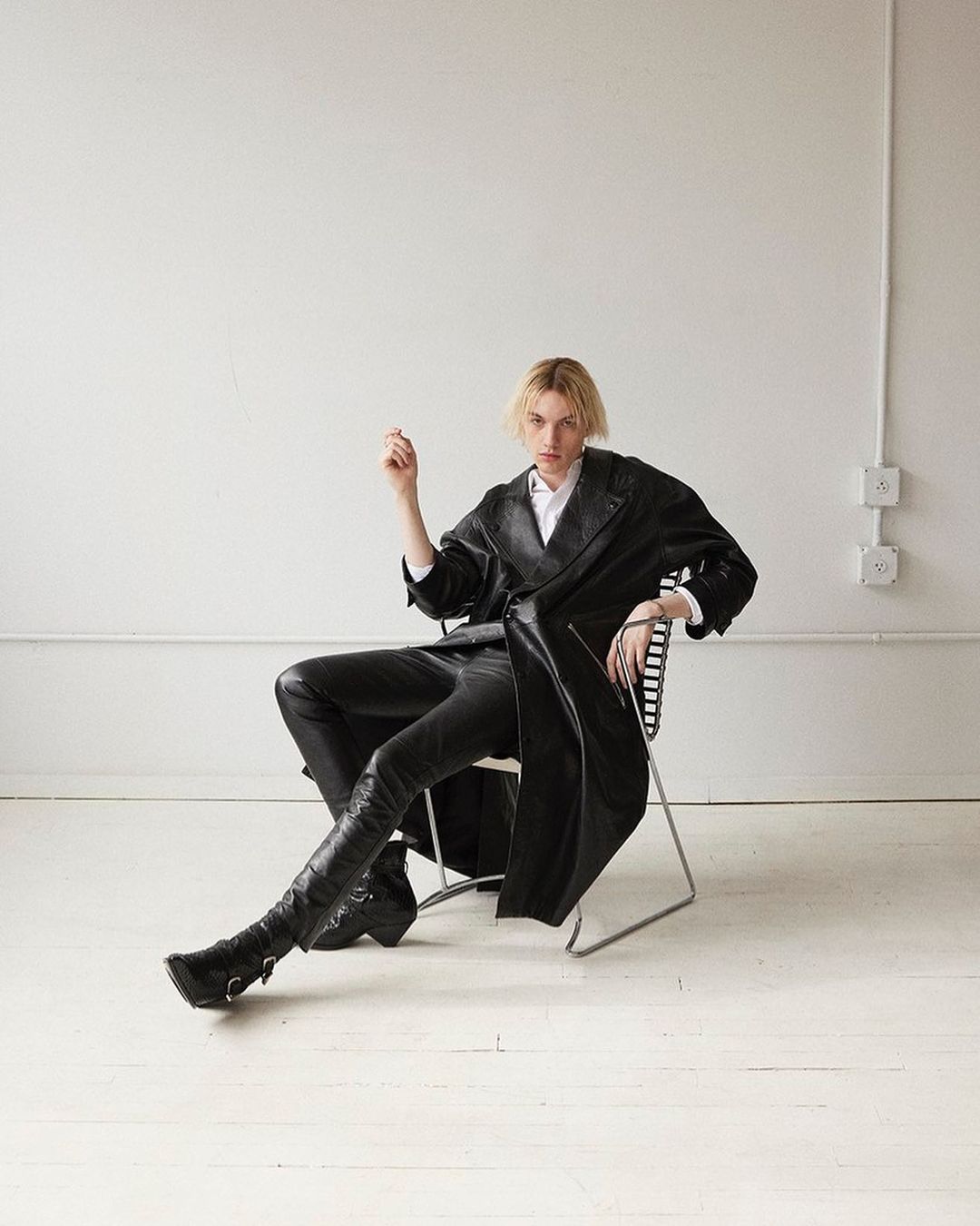 A person in a black leather-like trench coat, black leather-like pants, and black boots sitting on a chair in a photo studio.