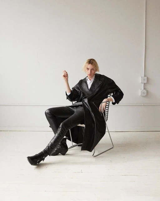 A person in a black leather-like trench coat, black leather-like pants, and black boots sitting on a chair in a photo studio.