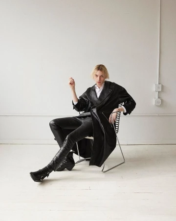 A person in a black leather-like trench coat, black leather-like pants, and black boots sitting on a chair in a photo studio.