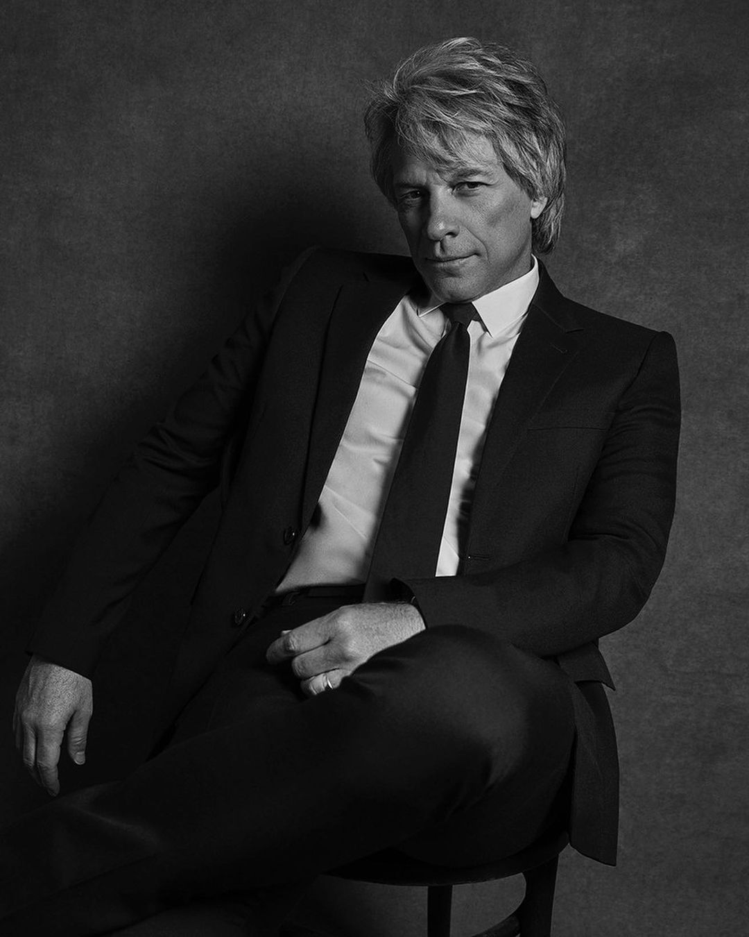 Black and white portrait of Jon Bon Jovi in a suit, seated on a stool against a dark backdrop, captured with a medium shot and eye-level angle.