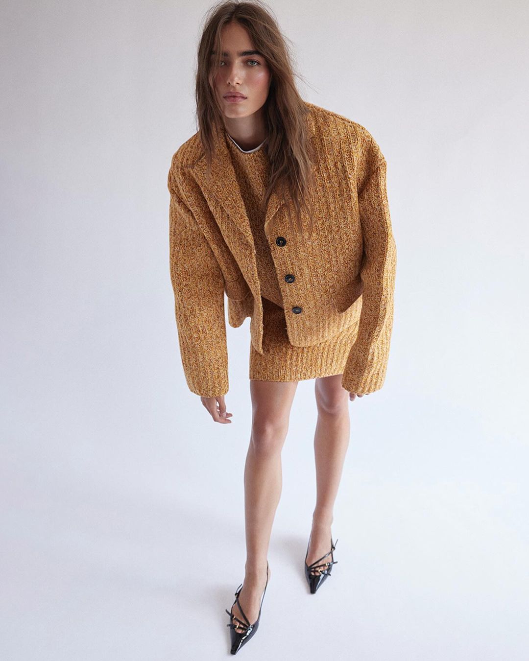 Woman in a mustard-colored knit skirt suit and black high heels poses against a white background in a fashion studio shot.