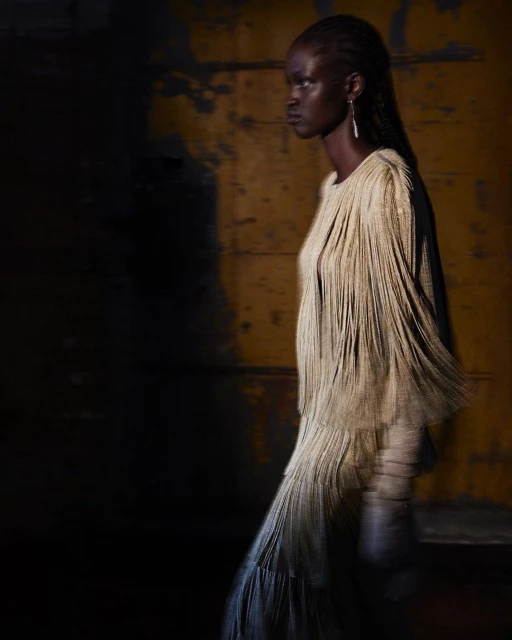 Fashion shot of an African woman in a cream-colored fringed dress and earrings against a mottled yellow background with dark shadows.