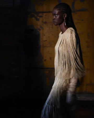 Fashion shot of an African woman in a cream-colored fringed dress and earrings against a mottled yellow background with dark shadows.