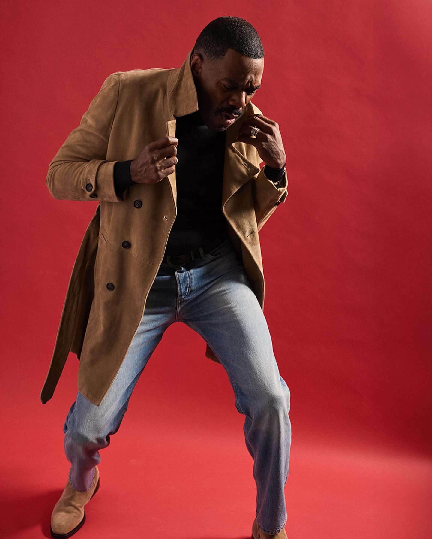 An African American man in a tan coat and blue jeans poses against a red background, showcasing a blend of casual and stylish fashion.