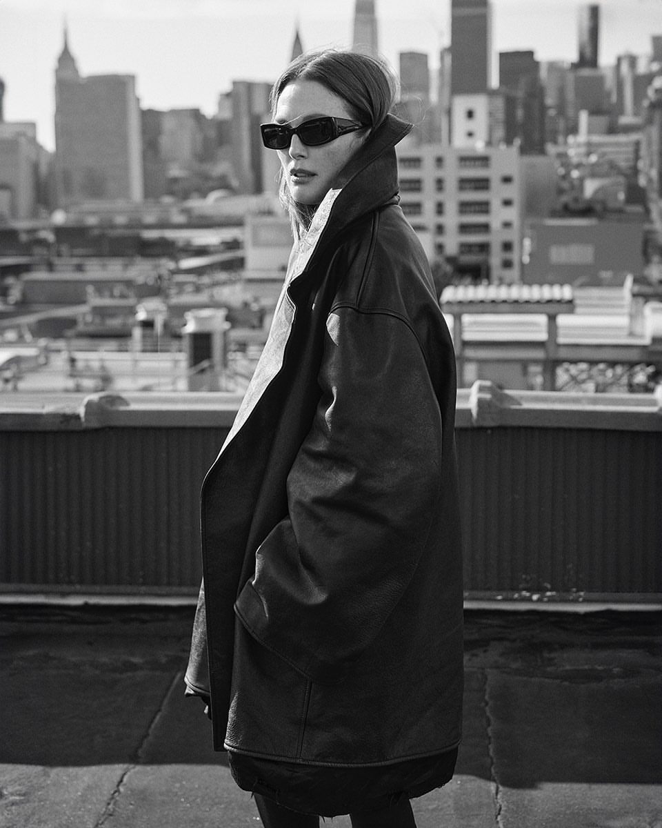 Black and white shot of woman in oversized leather jacket and sunglasses posing on a rooftop with city skyline in the background.