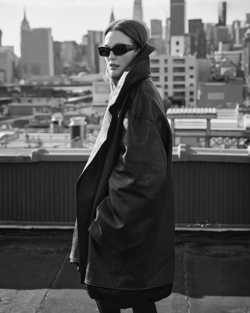 Black and white shot of woman in oversized leather jacket and sunglasses posing on a rooftop with city skyline in the background.