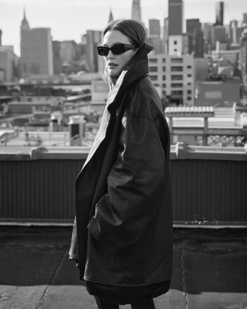 Black and white shot of woman in oversized leather jacket and sunglasses posing on a rooftop with city skyline in the background.