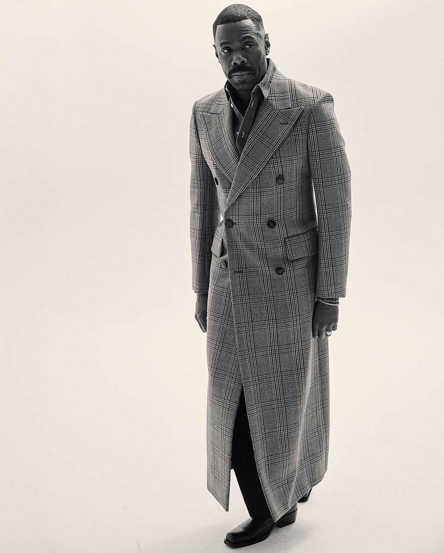 Portrait of an African-American man wearing a long plaid coat, button-up shirt and slacks, posing in a photo studio with soft lighting.