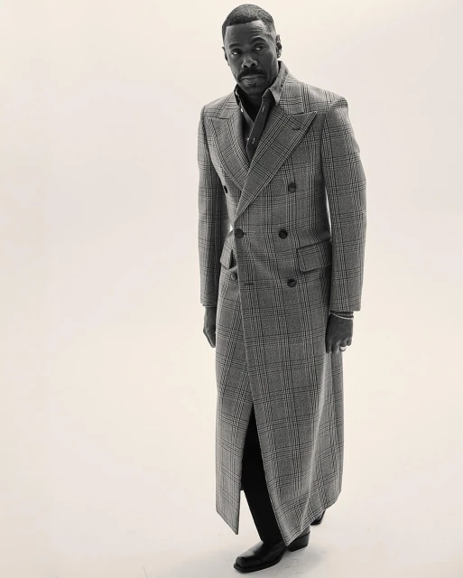 Portrait of an African-American man wearing a long plaid coat, button-up shirt and slacks, posing in a photo studio with soft lighting.