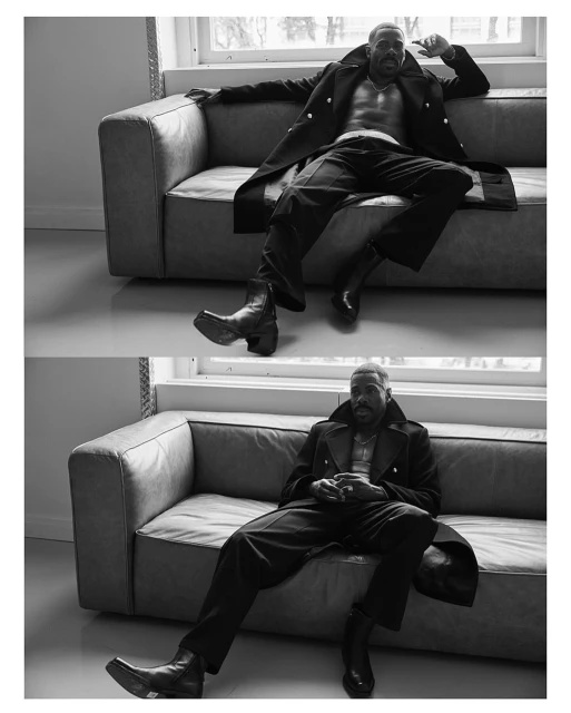 Black and white shot of an adult male model with a beard, wearing a coat and boots while posing on a couch in a studio.