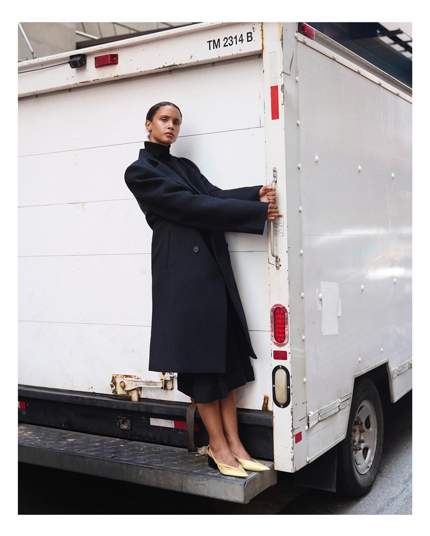 Woman in long black coat and yellow heels stands near the back of a white truck on a city street, fashion editorial photography.