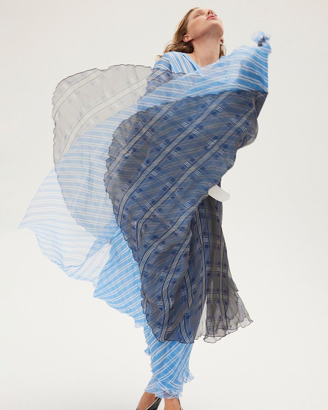Woman in flowing blue patterned dress poses against a white backdrop in a fashion editorial style full length shot.
