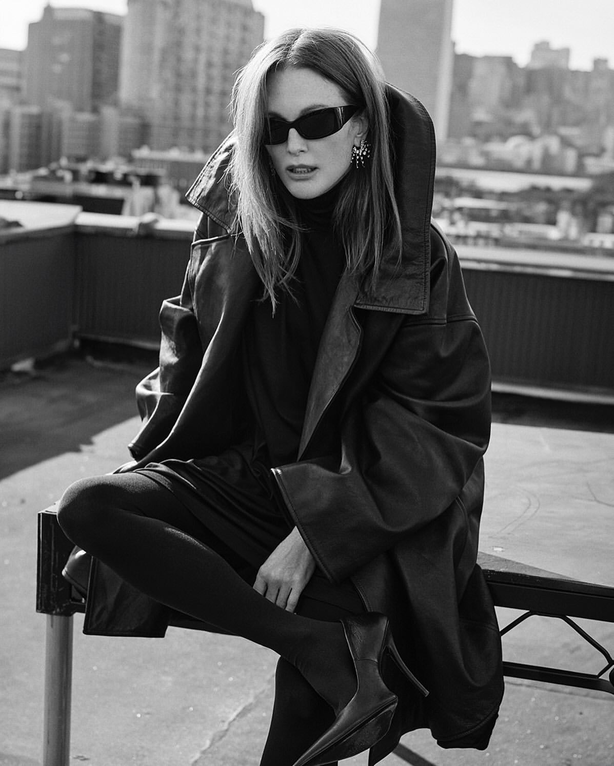 Black and white portrait of a woman in sunglasses, an oversized leather jacket, and pointed boots, posing on a rooftop in a city.