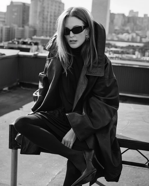 Black and white portrait of a woman in sunglasses, an oversized leather jacket, and pointed boots, posing on a rooftop in a city.
