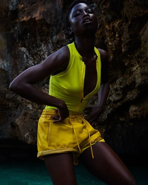 Dark-skinned adult woman in yellow workout attire poses in a rocky cave with turquoise water, looking up, hands on her hips.