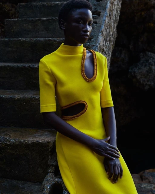 Woman in a yellow dress poses on stone steps outside. She has dark skin and short hair. The garment has cut-out details and interesting design.