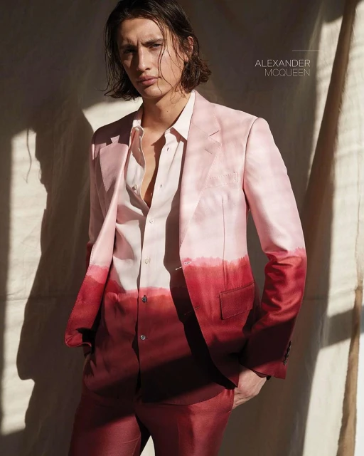 Male fashion model posing indoors in a pink and red dip-dye suit, wearing a light pink shirt with soft lighting and fabric backdrop.