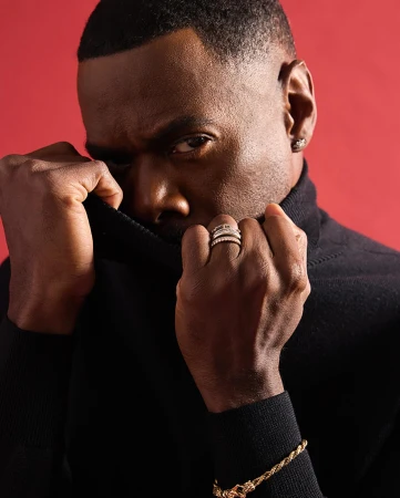 Close-up portrait of a Black man wearing a black turtleneck and rings, pulling up his collar against a red background.