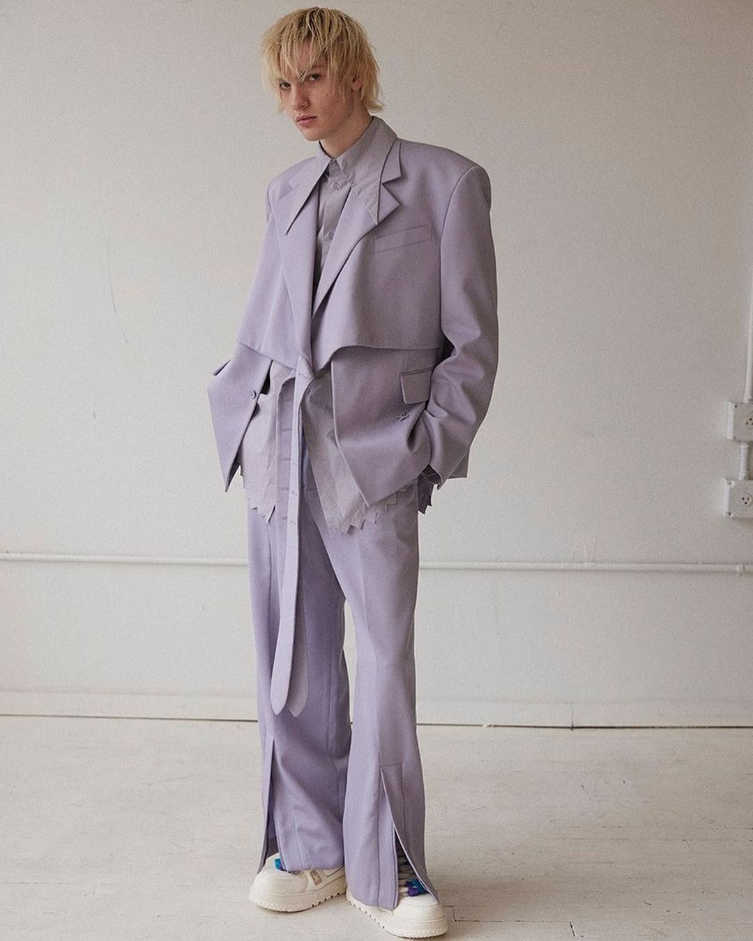 Full-length shot of androgynous model in lilac suit with jacket and tied belt, split-hem pants, and platform sneakers posing in studio.