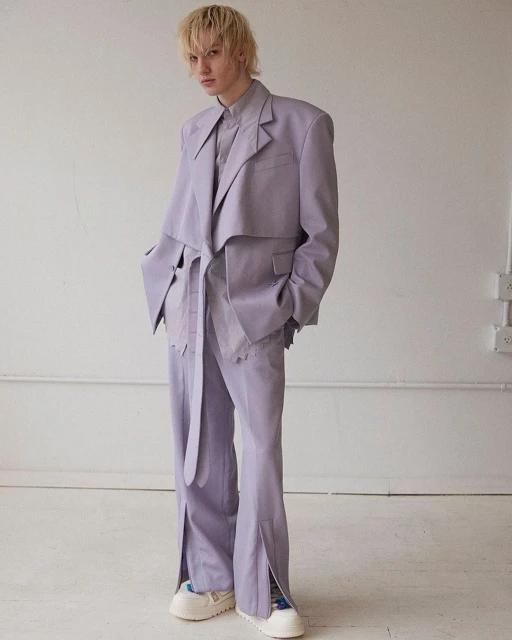 Full-length shot of androgynous model in lilac suit with jacket and tied belt, split-hem pants, and platform sneakers posing in studio.