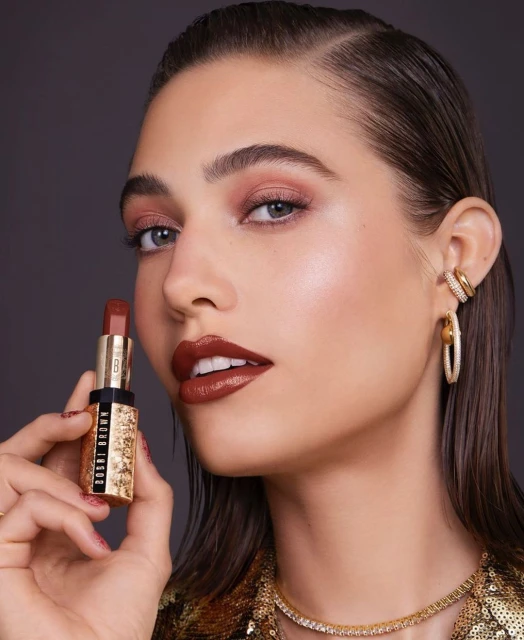 Close-up of an adult female holding Bobbi Brown lipstick, showcasing makeup and jewelry in a beauty-focused studio setting.