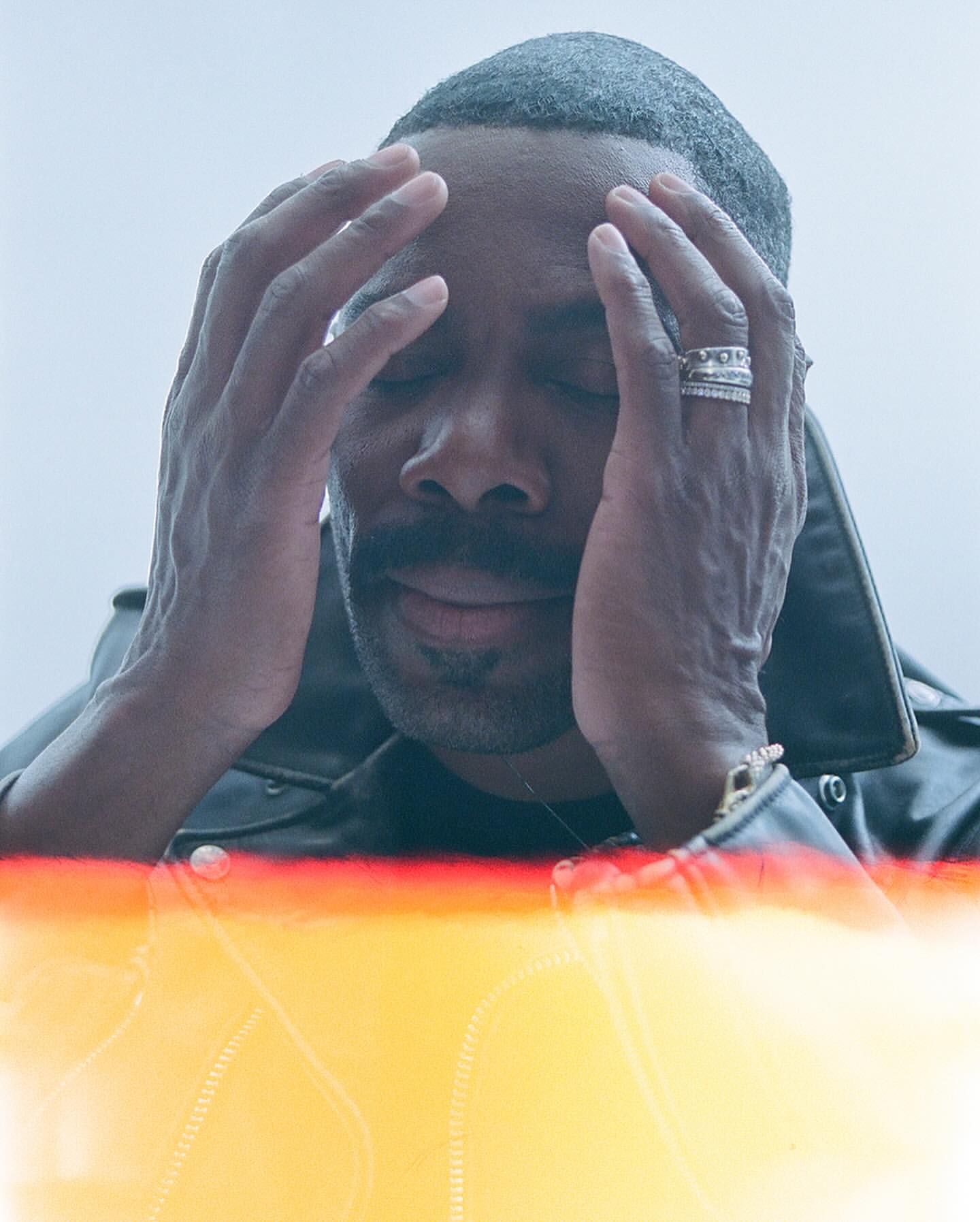 Close-up portrait of an adult man with dark hair, mustache, eyes closed, and hands on his face wearing a leather jacket indoors.
