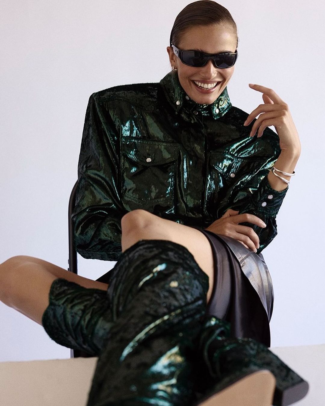 Smiling woman wearing dark sunglasses and a glossy emerald green jacket seated against a plain background