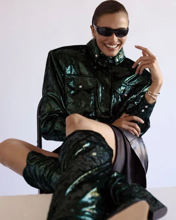 Smiling woman wearing dark sunglasses and a glossy emerald green jacket seated against a plain background