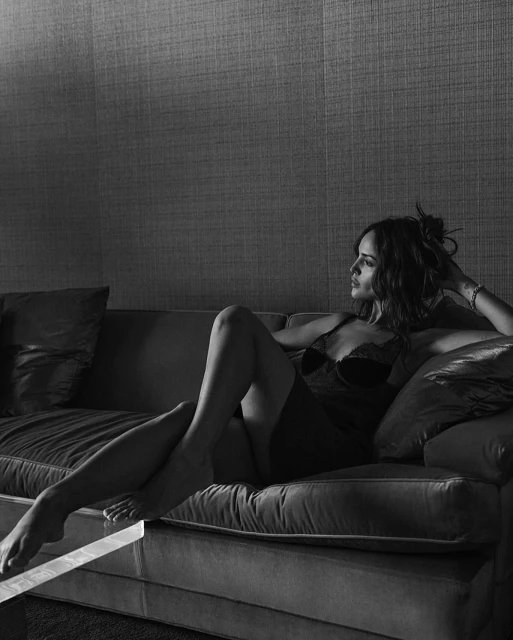 Monochrome portrait of an adult woman in lingerie reclining on a couch, her hand resting on her head as she gazes off into the distance.