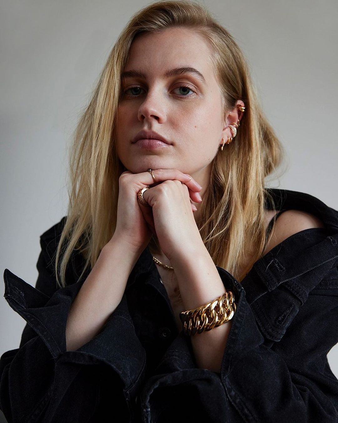 Portrait of a young, blond woman wearing a denim jacket and jewelry, gazing at the camera with her hands near her face.
