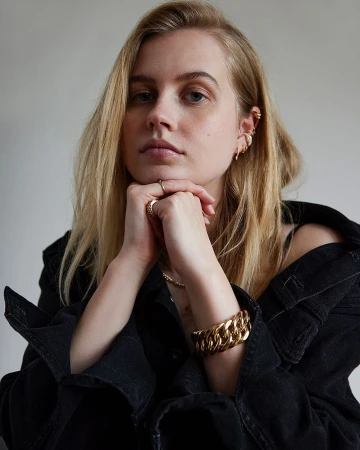 Portrait of a young, blond woman wearing a denim jacket and jewelry, gazing at the camera with her hands near her face.