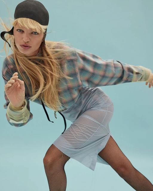 A blonde woman with a black cap poses in a studio wearing a plaid top, sheer skirt, and fishnet stockings, set against a light blue backdrop.