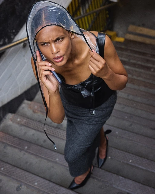 Woman in a black top, gray skirt, and clear rain bonnet on stairs using a smartphone, viewed from above. She is wearing black heels.