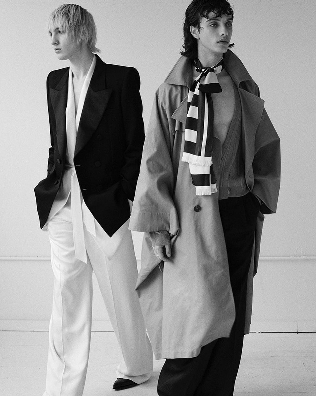 Black and white fashion shot features two slender male models in oversized blazers, trousers, and statement scarf against a plain backdrop.
