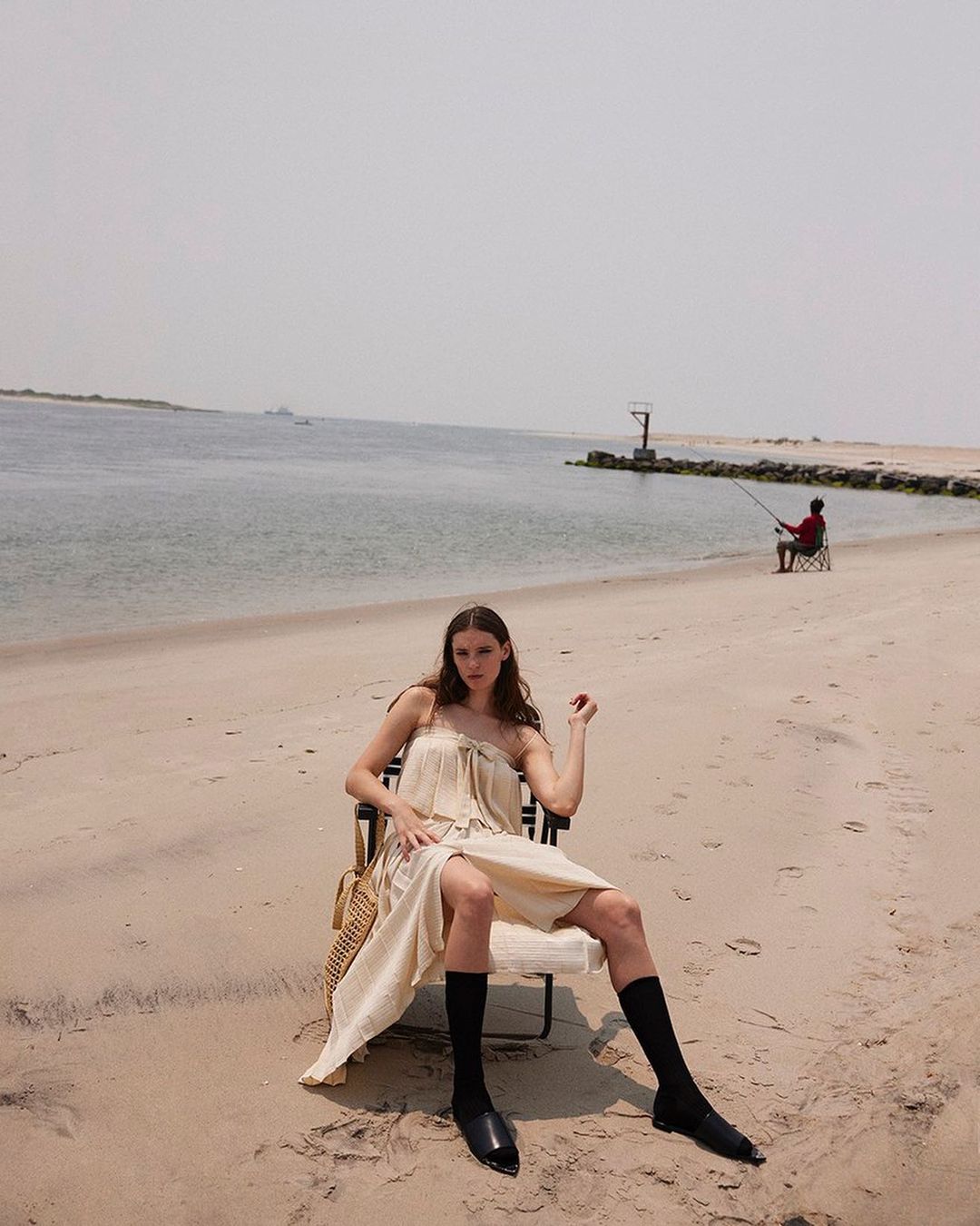 Fashion shot of a woman in a dress sitting in a chair on a beach with a fisherman in the background during daytime.