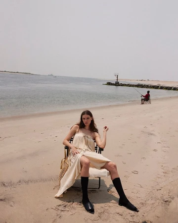 Fashion shot of a woman in a dress sitting in a chair on a beach with a fisherman in the background during daytime.