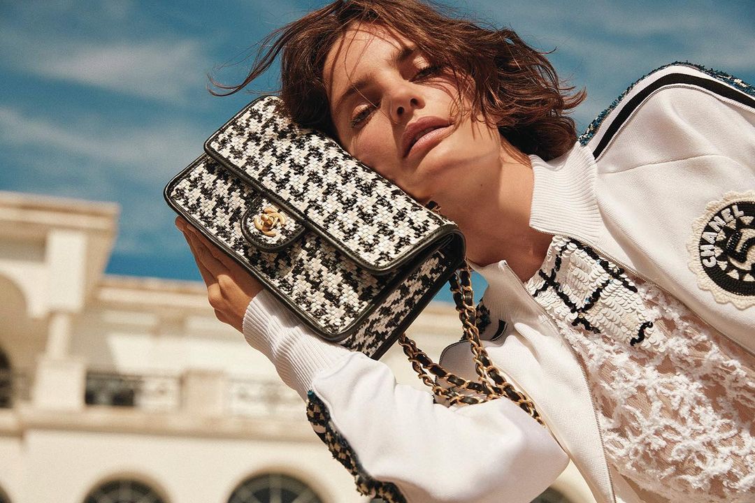 Close-up of a woman with short brown hair holding a patterned Chanel handbag with gold chain straps against a bright sky background.