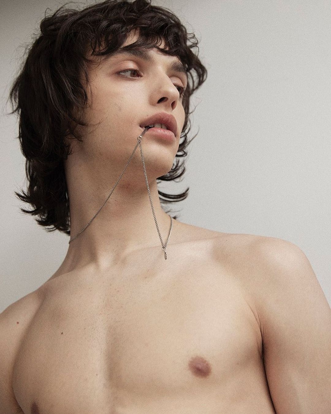 Portrait of a shirtless young man with dark curly hair and a necklace, looking up against a light-colored wall in a minimalist style.