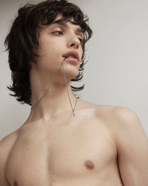 Portrait of a shirtless young man with dark curly hair and a necklace, looking up against a light-colored wall in a minimalist style.