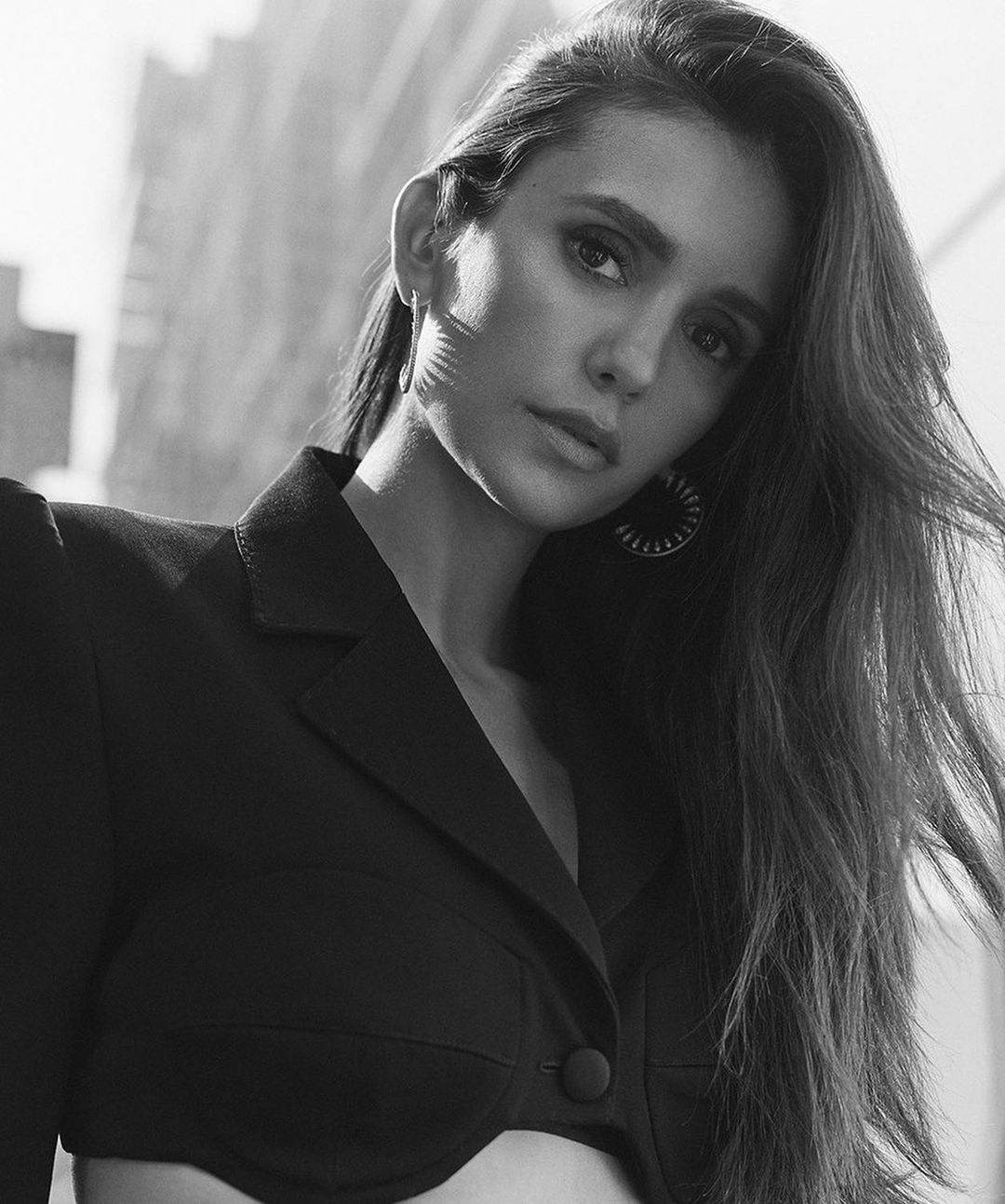 Black and white portrait of a woman with long brown hair wearing a jacket, set against a backdrop of blurred buildings.