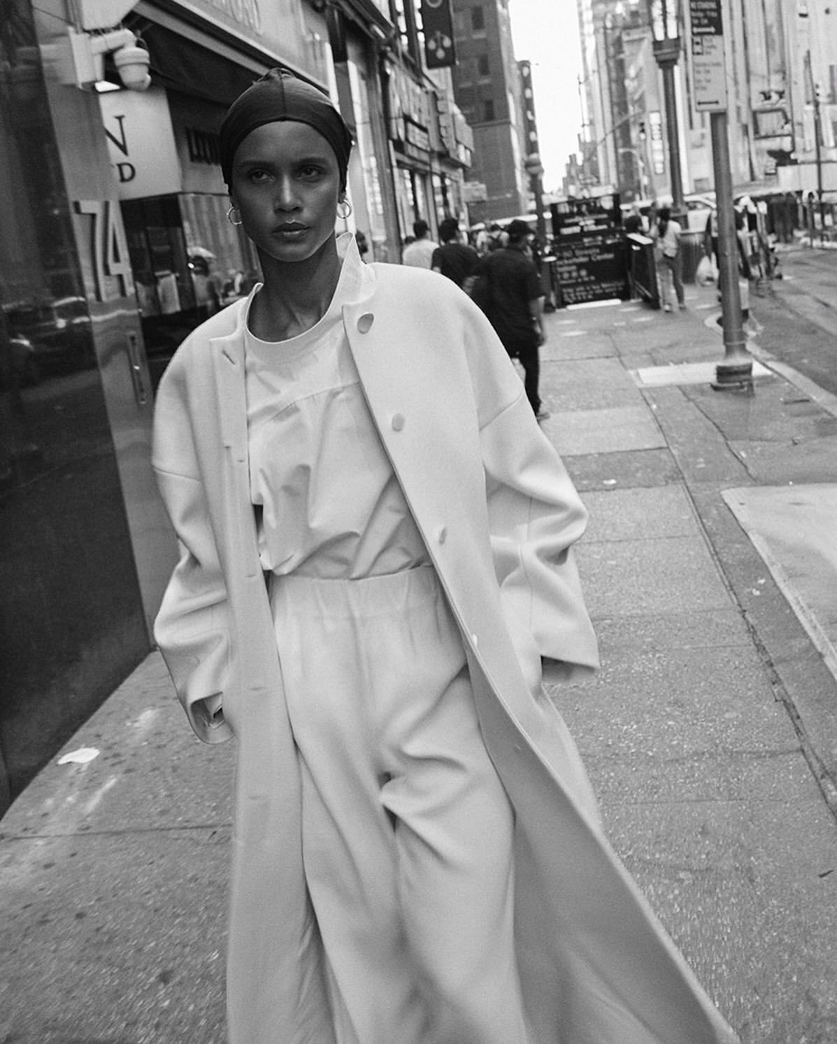 A black and white shot of a woman in a coat and head covering walking in an urban setting. The woman is the primary subject.