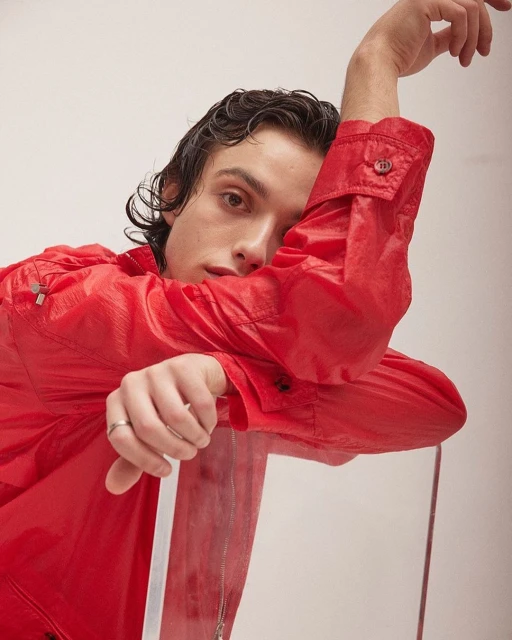 Portrait of a young adult male with dark hair, wearing a bright red semi-transparent jacket, leaning on a clear acrylic chair against a plain white background.