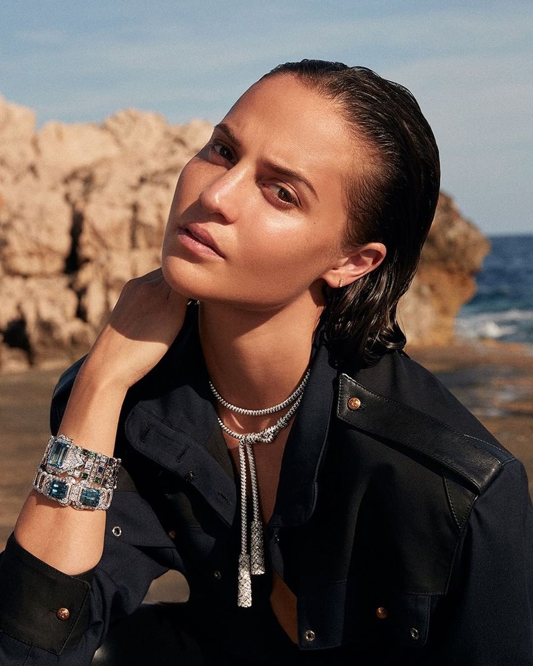 Woman with dark wet hair poses with bracelets and necklace in a fashion shot outdoors against a rocky shoreline on a sunny day.