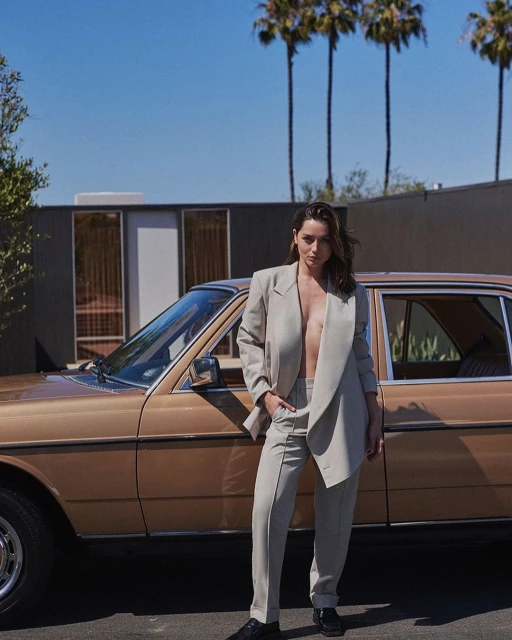 Adult woman in a beige pantsuit poses beside a vintage tan car on a sunny day with palm trees and a modern house in the background.