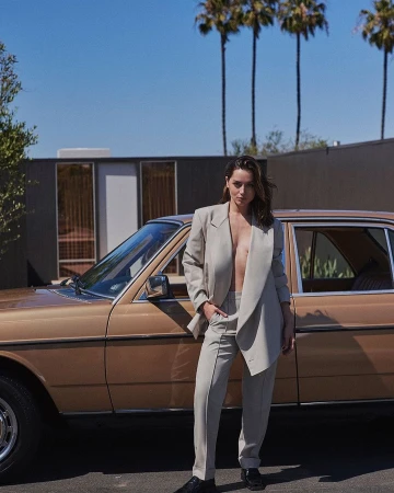 Adult woman in a beige pantsuit poses beside a vintage tan car on a sunny day with palm trees and a modern house in the background.