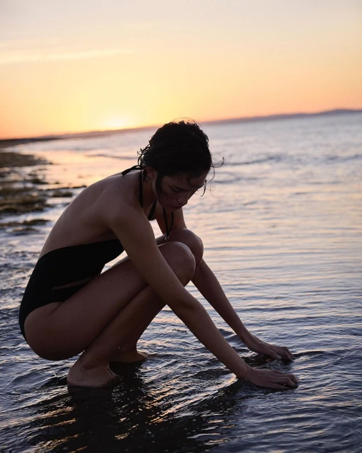 A woman in a black bathing suit squats in the ocean at sunset with her hands touching the water, creating a serene and picturesque scene.