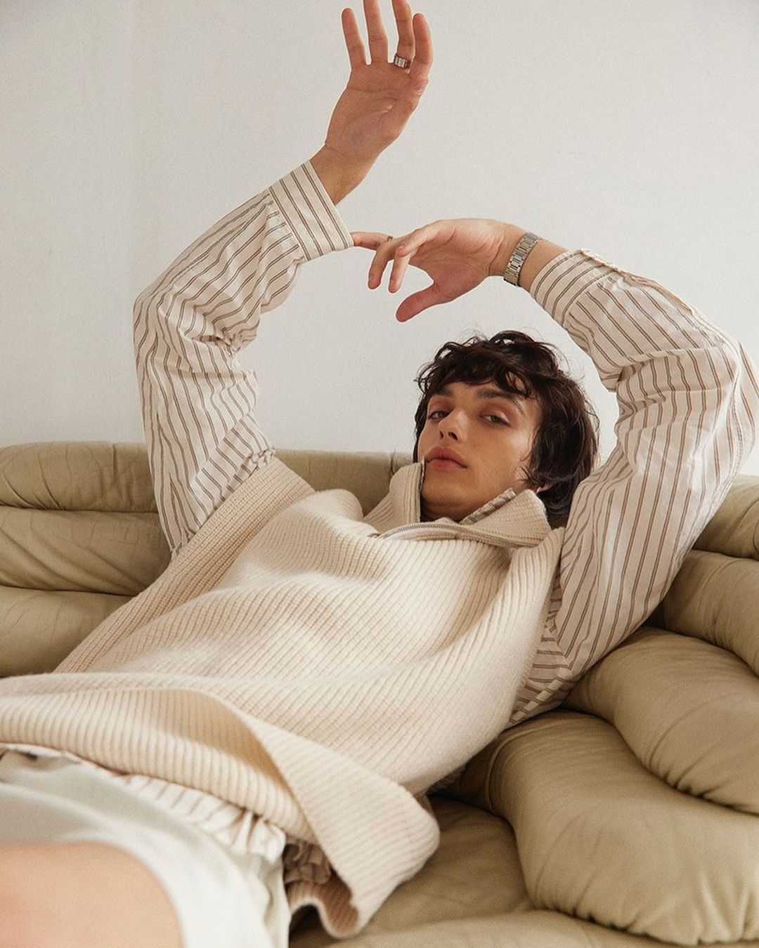 Adult male in a striped shirt and knit sweater vest poses while reclining on a leather couch in a relaxed setting. Indoor shot.