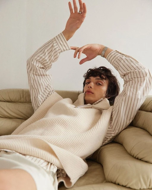 Adult male in a striped shirt and knit sweater vest poses while reclining on a leather couch in a relaxed setting. Indoor shot.